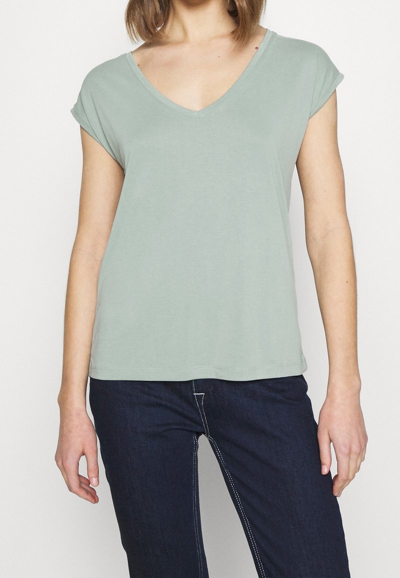 Femme portant un haut vert clair à manches courtes en V et un jean bleu foncé avec des coutures blanches, debout devant un fond uni.