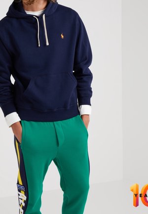 Persoon draagt een donkerblauwe hoodie met witte trekkoorden en groene sportbroek met gele en donkerblauwe strepen aan de zijkant, handen in de zakken.