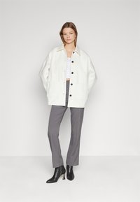 Bílá oversized bunda s velkými kapsami, černými knoflíky, spárovaná se šedými kalhotami na míru a černými kotníkovými botami. Hladká látka, minimalistický design.