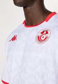 Balts sporta krekls ar sarkaniem akcentiem, ar teksturētu dizainu, sarkanu apkakli un apļveida logo kreisajā pusē. Kappa logo uz krūšu daļas.