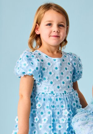 Jeune fille aux cheveux blonds mi-longs portant une robe bleu clair décorée de fleurs blanches et turquoises, debout sur un fond bleu uni.