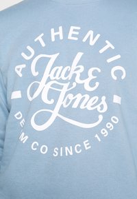 Světle modrá mikina s velkým bílým nápisem "AUTHENTIC Jack & Jones" a kulatým emblémem s textem "DENIM CO SINCE 1990".
