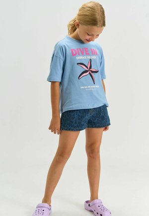 Vaaleansininen grafiikkapaita, jossa vaaleanpunainen meritähden kuvio, yhdistetty tummansinisiin leopardikuvioisiin shortseihin ja purppuranvärisiin puukenkäisiin. Arkiasu.