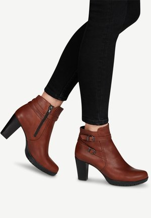 Botas de cuero marrón hasta el tobillo con tacón alto, que cuentan con una cremallera lateral y dos detalles de hebilla en el tobillo para un toque adicional.