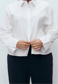 Chemise blanche à boutons avec col, manches longues et détails aux poignets. Tissu lisse, coupe ajustée, portée sur un pantalon foncé.