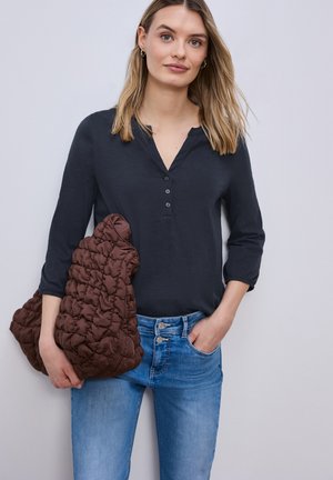 Kobieta o prostych blond włosach, ubrana w granatową koszulkę henley i niebieskie dżinsy, trzyma w jednej ręce dużą brązową pikowaną kurtkę.