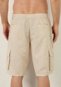 Pantaloni scurți cargo beige, până la genunchi, cu buzunare tardive la spate și buzunare laterale cu clapă, purtați de o persoană fără cămașă în fața unei pereți deschisi la culoare.