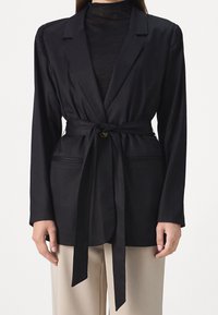 Schwarze Blazer mit Gürtel aus glattem Stoff und tailliertem Schnitt, mit einem Ein-Knopf-Verschluss und Seitentaschen.