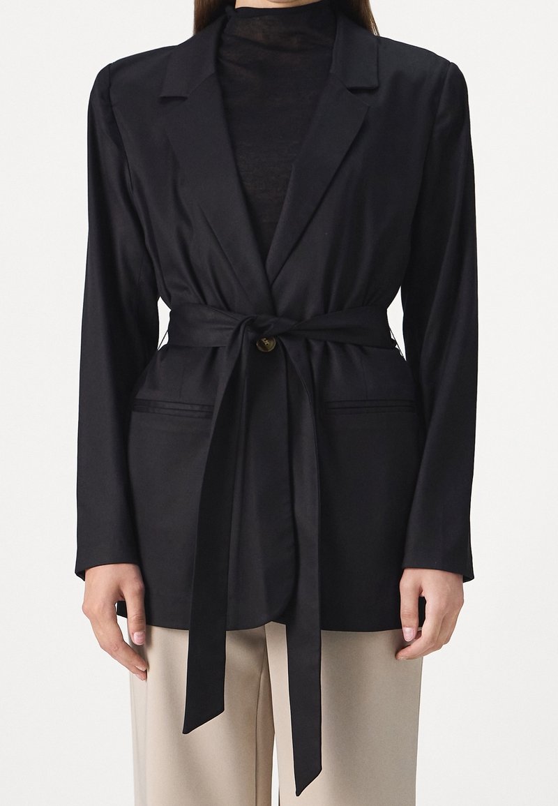 Schwarze Blazer mit Gürtel aus glattem Stoff und tailliertem Schnitt, mit einem Ein-Knopf-Verschluss und Seitentaschen.