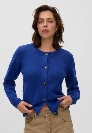 Jeune femme aux cheveux bruns portant un pull bleu boutonné et un pantalon beige, ajustant le pull près de la taille.
