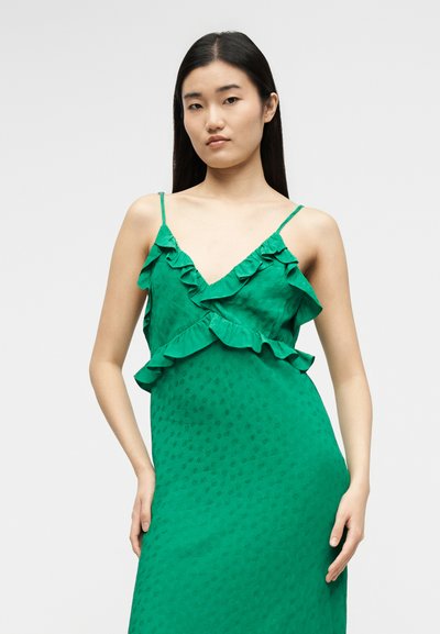 Γυναίκα με μακριά σκουρόχρωμα μαλλιά, που φοράει πράσινο αμάνικο φόρεμα με βολάν κατά μήκος της λαιμόκοψης και της μέσης, στέκεται μπροστά σε απλό φωτεινό φόντο.