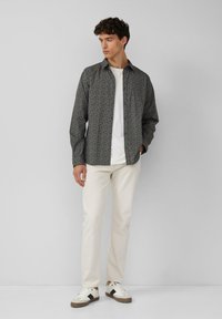 Overhemd met knopen in een donkere, gedessineerde stof, een wit T-shirt met ronde hals eronder, gecombineerd met lichte broek en witte sneakers.