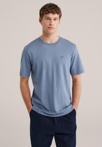 Jonge man met krullend haar, gekleed in een lichtblauw T-shirt met korte mouwen en een klein wit handlogo, gecombineerd met donkerblauwe broek, handen in de zakken.