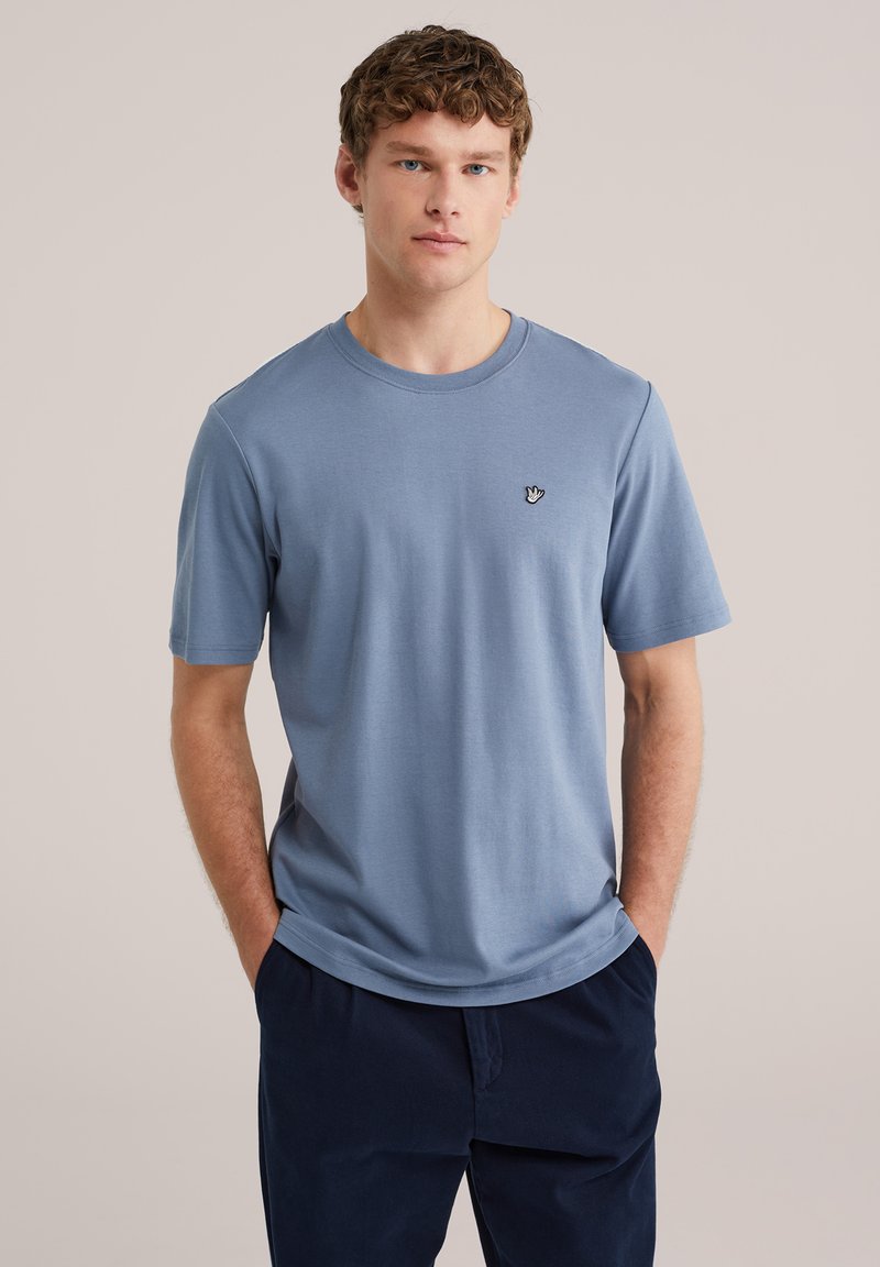 Jonge man met krullend haar, gekleed in een lichtblauw T-shirt met korte mouwen en een klein wit handlogo, gecombineerd met donkerblauwe broek, handen in de zakken.