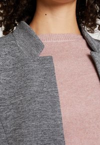 Harmaan tekstuurisen bleiserin kaulus vaaleanpunaisen ribbineuleen päällä, esitellen kontrastisia värejä ja materiaaleja lähikuvassa.