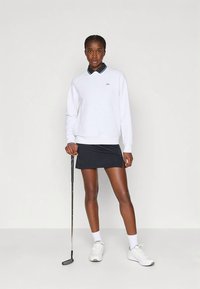 Nő fehér hosszú ujjú pólóban, fekete szoknyában, fehér sportcipővel, golflabdával a kezében, sima világos háttér előtt áll.