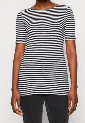 Persoon draagt een zwart-wit horizontaal gestreept shirt met korte mouwen en een zwarte broek tegen een effen lichte achtergrond.
