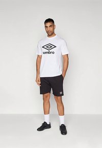 Baltas medvilninis marškinėlis su juodu "Umbro" logotipu, derinamas su juodais šortais ir juodais sportiniais bateliais, su baltais kojinaitėmis ir minimalistiniu dizainu.