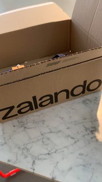 Brūna kartona kaste ar lieliem melniem burtiem uzrakstītu "zalando". Iekšpuse daļēji redzama, bet saturs ir neskaidrs. Virsma ir ar marmora tekstūru.