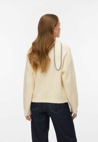 Krēmkrāsas džemperis ar ribotu tekstūru, noapaļotu kakla izgriezumu un pelēku apmali aizmugurē. Kombinēts ar tumšām džinsām, skatīts no aizmugures.