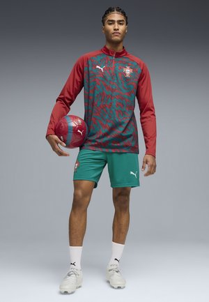 Nuori mies seisoo ja pitää kädessään puna-turkoosia jalkapalloa, pukeutuneena Portugalin jalkapallopaitaan, jossa on Puma-logot, harmaan taustan edessä.