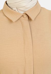 Camisa beige de botones con cuello estructurado, hecha de una tela suave, con una textura lisa y un diseño minimalista.
