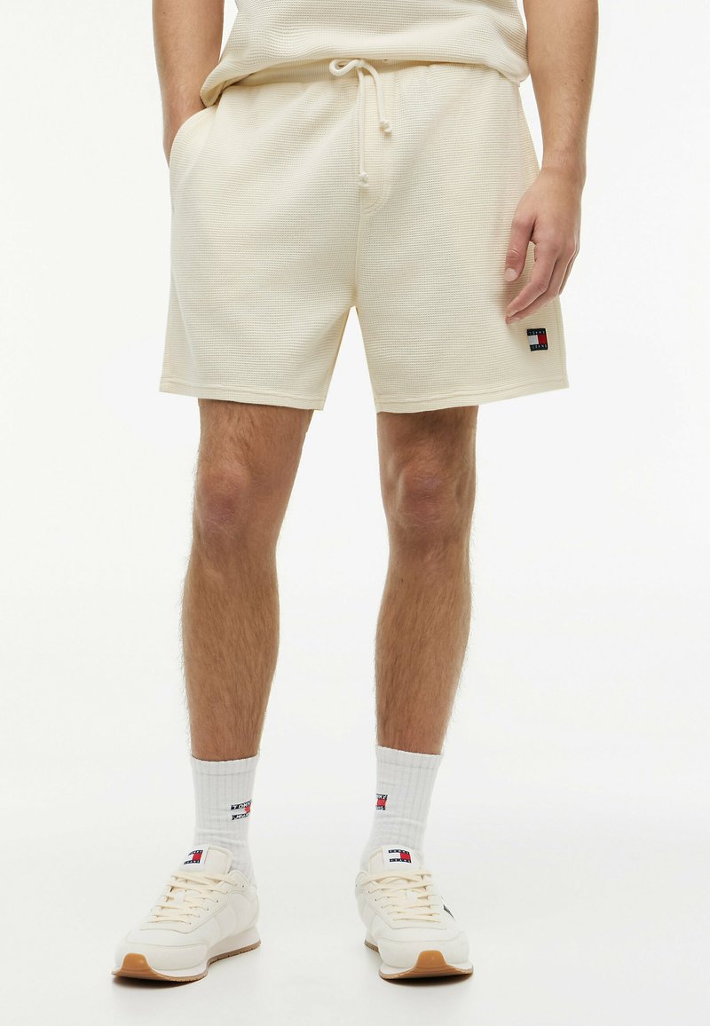 Homme portant un short texturé beige clair avec poches latérales, des chaussettes blanches de marque et des baskets beige clair, debout les mains dans les poches.