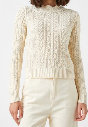 Femme portant un pull en maille torsadée de couleur crème à manches longues et poignets côtelés, associé à un pantalon crème clair taille haute.