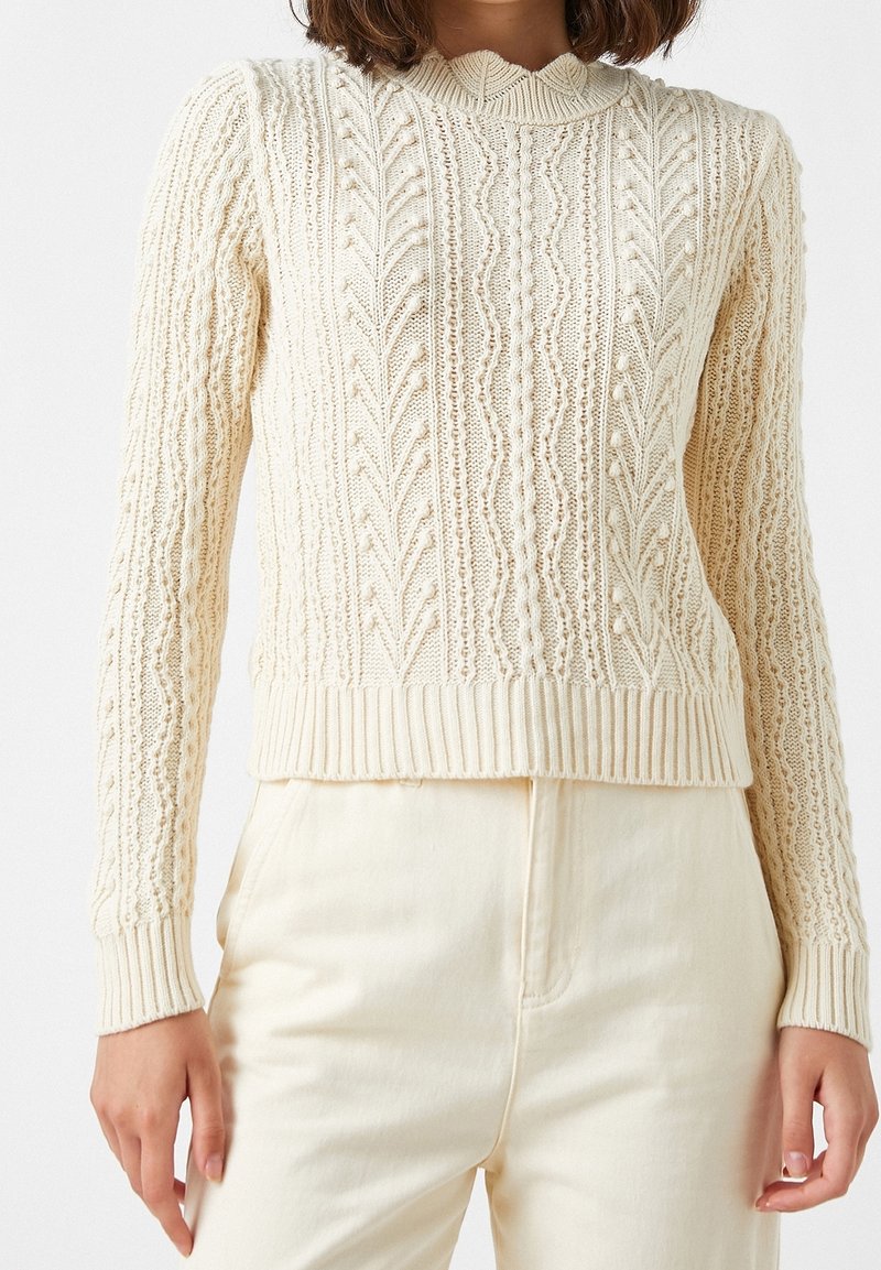 Femme portant un pull en maille torsadée de couleur crème à manches longues et poignets côtelés, associé à un pantalon crème clair taille haute.