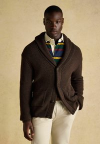 Barna bordázott kötött kardigán sálgallérral, nagy gombokkal és két elülső zsebbel, csíkos pólóing fölött viselve.