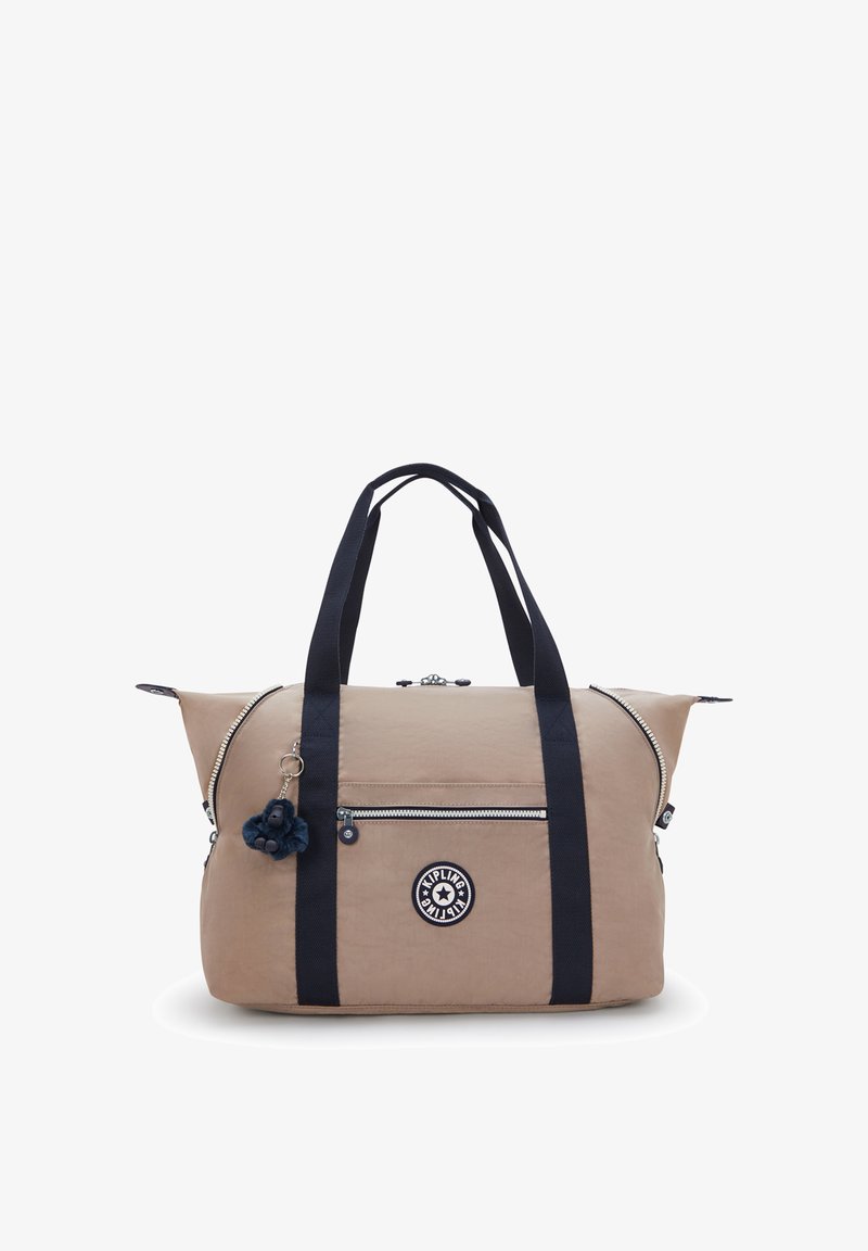 Beige stofduffeltaske med marineblå håndtag, lynlåslukninger, forlommer og en mørkeblå pompom-vedhæng. Har en logo-patch.