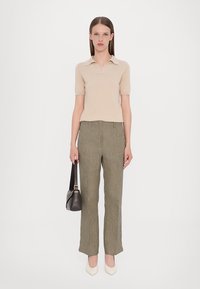 Polo shirt beige a maniche corte abbinato a pantaloni verdi oliva con motivo. Il modello tiene una borsa nera e indossa scarpe bianche a punta.