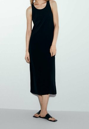 Femme portant une robe midi noire sans manches et des sandales noires à enfiler, debout devant un fond clair uni.