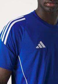 Blå idretts skjorte med hvite striper på skuldrene, med en grå Adidas-logo. Stoffet ser glatt ut med en tettsittende design.