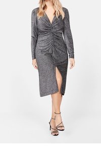 Vestido gris metálico con mangas largas, detalle fruncido en el costado, escote en V y abertura hasta el muslo, combinado con zapatos de tacón alto negros con tiras.