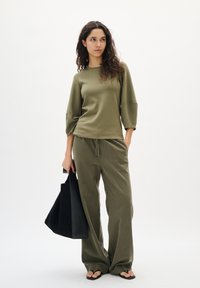 Olivgrünes Stricktop mit Dreiviertelärmeln, kombiniert mit passenden weit geschnittenen Hosen. Eine schwarze Tote-Tasche und Flip-Flops vervollständigen das Outfit.