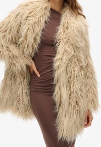 Faux fur-kappa i beige, med en lång, shaggy textur och öppen front, matchad med en kroppsnära brun klänning under.