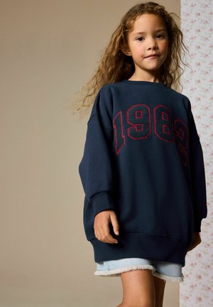 Marineblå sweatshirt med rød syning, der har tallet 1982, løs pasform, ribstrikkede ærmegab, kombineret med lyse denimshorts med frynset kant.