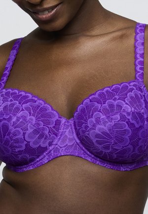 Woman wearing a purple lace underwire bra with scalloped edges, showing a close-up of the chest and shoulder area.