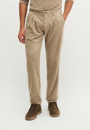 Mann trägt beige Cordhosen mit Bundfalten, kombiniert mit braunen Schnürschuhen vor einem einfarbigen hellen Hintergrund.
