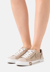 Sneakers beige in suede strutturato con lacci bianchi, una cerniera laterale e un dettaglio logo marrone a contrasto sul tallone.