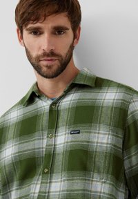 Zielono-biała koszula flanelowa w kratę z kołnierzykiem na guziki i małą niebieską metką. Charakteryzuje się miękką fakturą i casualowym designem.