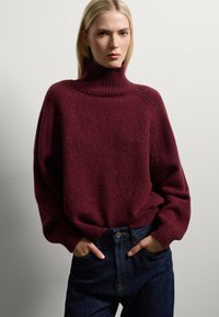 Suéter de punto burdeos con un alto cuello acanalado, corte oversized, hilo texturizado y hombros caídos, combinado con jeans de mezclilla azul oscuro.