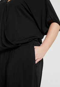 Personne portant des vêtements noirs amples à manches courtes et taille élastique, main posée négligemment dans la poche latérale sur fond clair.