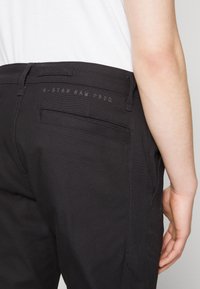 Zwarte stoffen broek met een gestructureerde afwerking, voorzien van achterzakken en het geëmbosste logo "G-STAR RAW PROD." op de tailleband.