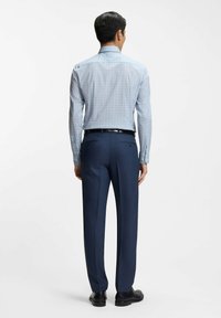 Man draagt een lichtblauw overhemd met patroon, ingestopt in donkerblauwe nette broek met zwarte riem en zwarte nette schoenen, staand met de rug naar de camera.