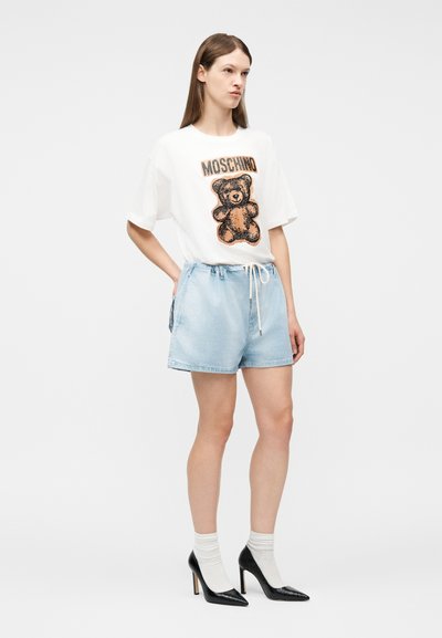 Γυναίκα που φοράει λευκό t-shirt Moschino με γραφικό αρκουδάκι, ανοιχτό μπλε τζιν σορτς, λευκές κάλτσες και μαύρα ψηλά τακούνια, στέκεται με το χέρι στη μέση.