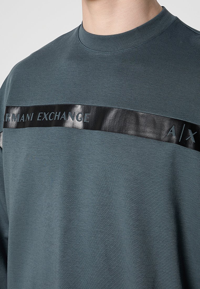 Nahaufnahme einer Person, die einen dunkelblauen Sweatshirt mit einem glänzenden schwarzen horizontalen Streifen und dem Text "ARMANI EXCHANGE A/X" über der Brust trägt.