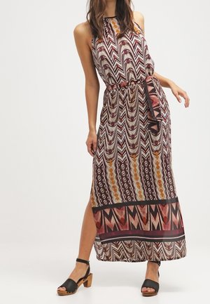Femme portant une robe maxi sans manches à motifs géométriques et tribaux, taille ceinturée, fente latérale et sandales noires à talon bloc et orteils ouverts.