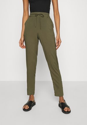 Frau trägt olivgrüne Hose mit Kordelzug, schwarzes ärmelloses Oberteil und schwarze Sandalen, steht vor einem einfachen hellen Hintergrund.